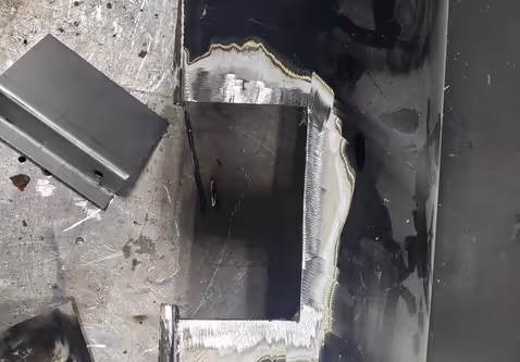 A patch panel repair in progress on a black car fender with the damaged section cut out and prepped for welding.