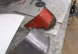 A newly fabricated, curved metal panel sits next to the old, rusted red part it was designed to replace, highlighting precision metal duplication.