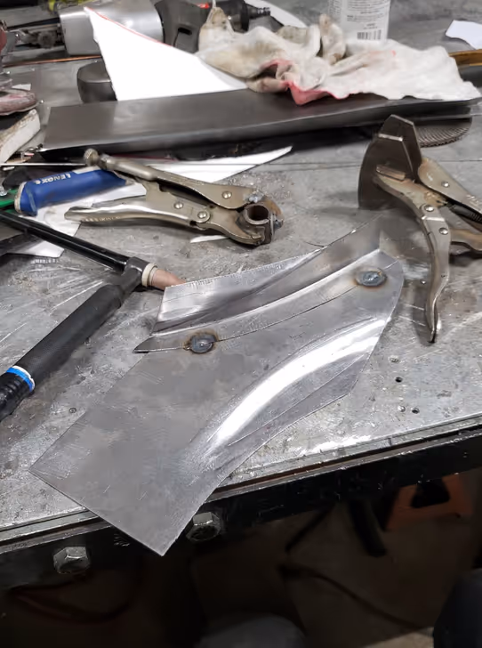 A custom-shaped steel panel with spot welds, held by clamps on a metal workbench next to a TIG welding torch.