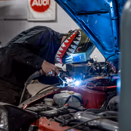 Fabricator welding on red vehicle with custom shark helmet